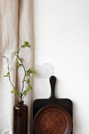 Photo for Brown vase with green tree branch, wooden board, ceramic clay plate, beige linen curtain, empty white wall background. Authentic sustainable home kitchen interior decor, natural organic beverage. - Royalty Free Image