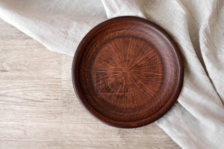 Téléchargez les photos : Assiette d'argile brune vide sur table en bois beige neutre et nappe en lin, vue du dessus, maquette vierge pour le placement de produits alimentaires. - en image libre de droit