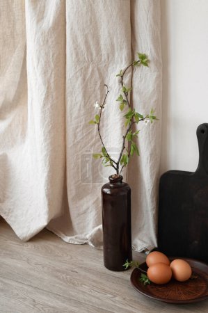 Photo for Elegant sustainable rustic Easter kitchen interior, natural color eggs on brown ceramic plate, vase with green tree branch on table, beige linen curtain, empty white wall with soft sunlight shadows - Royalty Free Image