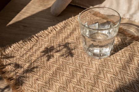 Téléchargez les photos : Verre avec de l'eau sur la table avec fond de tapis de jute beige sable avec des ombres de soleil abstraites florales naturelles, concept de soins de santé style de vie d'été, espace de copie. - en image libre de droit