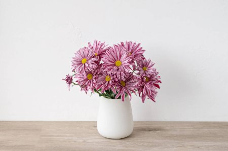 Téléchargez les photos : Bouquet de fleurs rose marguerite en vase céramique blanche sur table en bois beige neutre, fond mural blanc, décoration intérieure minimaliste esthétique florale, lumière naturelle douce. - en image libre de droit