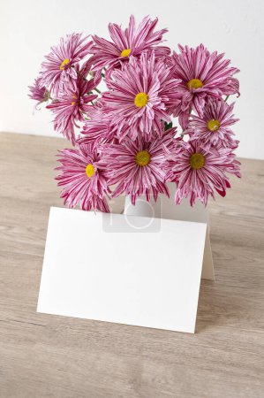 Aesthetic botanical spring holiday greeting card or wedding invitation design template, blank paper card mockup, vase with pink daisy flowers bouquet on beige wooden table background, copy space.
