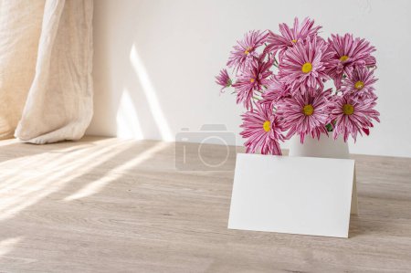 Empty paper card mockup, vase with pink flowers bouquet on beige wooden table, white wall and linen curtain background with natural sunlight shadows. Aesthetic greeting postcard or invitation template