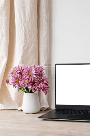 Téléchargez les photos : Esthétique espace de travail de bureau à domicile, ordinateur portable écran blanc maquette, vase avec bouquet de fleurs de chrysanthème rose sur table en bois beige, mur blanc vide et fond rideau de lin. - en image libre de droit