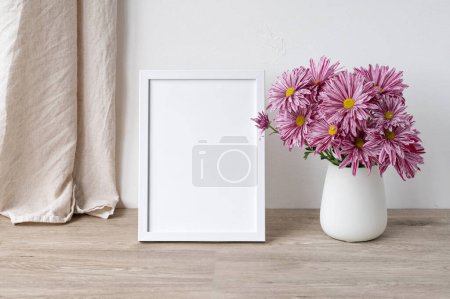 Foto de Marco de fotos blanco vacío o maqueta de póster, florero con ramo de flores rosadas en la mesa de madera beige claro, cortina textil de lino y fondo de pared blanco, bodegón estético en estilo nórdico. - Imagen libre de derechos