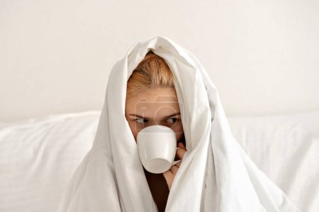 Foto de Chica adolescente soñolienta envuelta en manta de algodón blanco bebiendo café en la cama, hora de la mañana, concepto de despertar duro. - Imagen libre de derechos