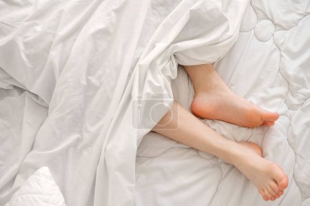 Foto de Pies desnudos en la cama bajo una manta de algodón blanco, buen sueño, concepto de ropa de cama para dormir confort, dormitorio nórdico minimalista. - Imagen libre de derechos