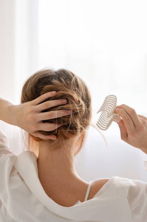 Photo for Fair haired girl in white shirt making messy hair bun, clipping with claw hair clip indoor with soft natural light, easy fashion woman hairstyle, daily beauty routine at home, lifestyle, back view. - Royalty Free Image