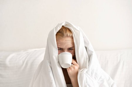 Téléchargez les photos : Café du matin, concept de réveil matinal difficile, jeune adolescente couverte d'une couverture blanche assise sur le lit dans une chambre lumineuse neutre, buvant du café dans une tasse en céramique blanche. - en image libre de droit