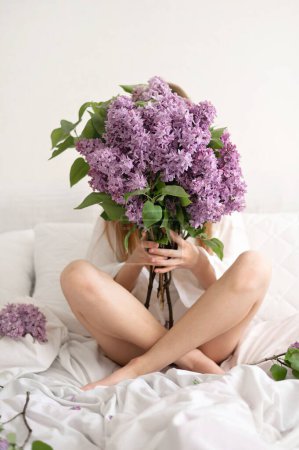 Téléchargez les photos : Young teenage girl sitting on bed with legs crossed, holding big lilac flower bouquet, hiding face behind, romantic femininity and self care concept. - en image libre de droit