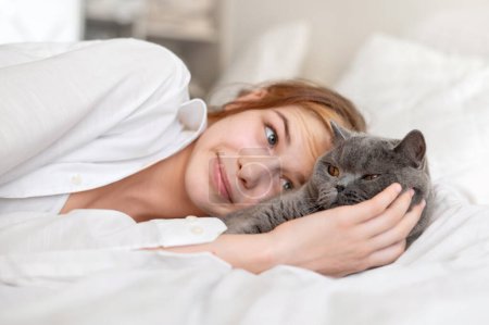 Foto de Teenage girl stroking gray cat laying in bed, pets care and love. - Imagen libre de derechos