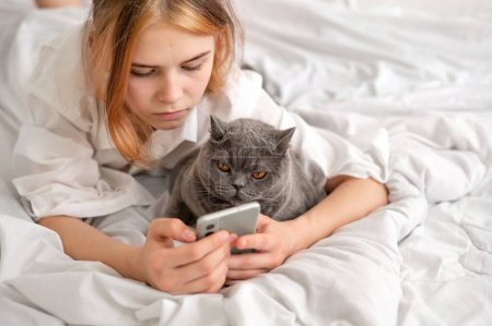 Teenager daily habits, girl using mobile phone, scrolling social media laying on bed together with her cat, child and pet friendship.