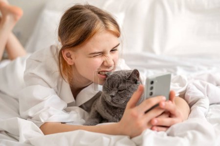 Lustiges Teenager-Mädchen mit Handy, umarmt graue Katze, beißt Katze Ohr, Kind verbringt Zeit mit Haustier und sozialen Medien zu Hause.