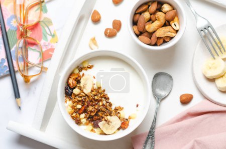 Téléchargez les photos : Petit déjeuner sain avec yaourt, granola, noix et banane sur table blanche. - en image libre de droit