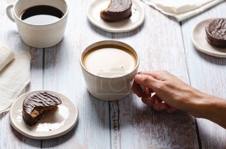 Téléchargez les photos : Fille boire du café et manger des alfajores au chocolat sur une table en bois. - en image libre de droit