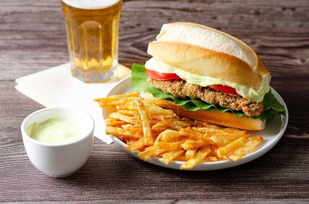 Téléchargez les photos : Sandwich milanesa avec tranches de tomate, laitue et mayonnaise et frites dans une assiette blanche, un bol avec mayonnaise et un verre de bière sur fond de bois. - en image libre de droit