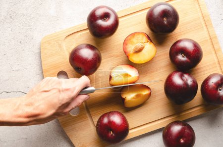 A woman's hand cutting a plum with a knife on a wooden cutting board an grey background.