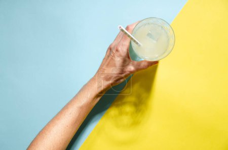 A left hand holding a glass of lemonade with a straw on blue and yellow background with copy space. 