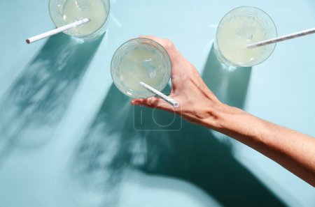 Three glasses of lemonade with straw and a right hand holding one of them on blue background. 