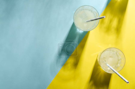 Zwei Gläser Limonade mit einem Strohhalm und der Schatten eines anderen Glases auf hellblauem und gelbem Hintergrund mit Kopierraum. 