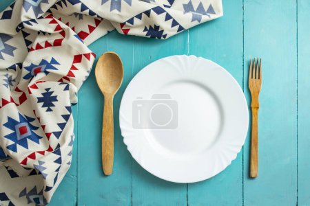 Leerer weißer Teller mit Holzbesteck und Serviette mit rumänischen ethnischen Motiven auf blauen Holzbrettern, rustikaler Hintergrund. 