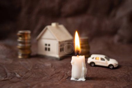 Kerze brennend mit Münzen, Miniaturhaus und Auto im Hintergrund, auf braunem Leder, Reichtum im Angesicht des Todes