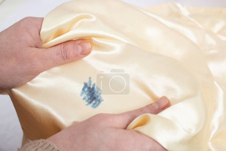 Woman hands stretching golden satin fabric with a blue ink stain, soft focus close up