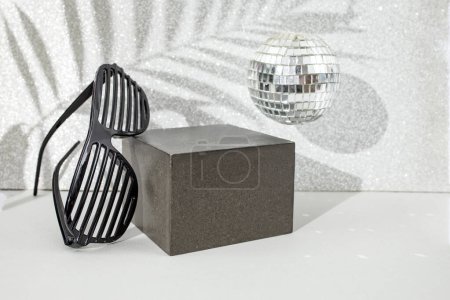 Gray backdrop with cube , party grate shades, disco mirror ball and palm leaf shadows on a silver glitter wall. Empty stage on grey background for product  