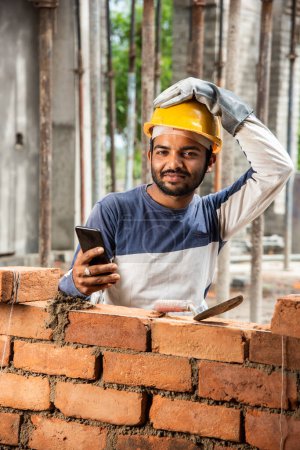 Téléchargez les photos : Joyeux sourire jeune travailleur indien de la construction occupé sur téléphone mobile sur le lieu de travail - en image libre de droit