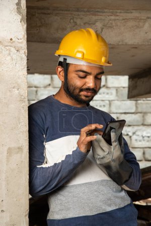 Téléchargez les photos : Joyeux sourire jeune travailleur indien de la construction occupé sur téléphone mobile sur le lieu de travail - en image libre de droit