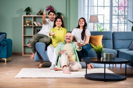 Téléchargez les photos : Famille indienne de quatre sur canapé, absorbé par regarder la télévision, changer de chaînes avec télécommande - en image libre de droit