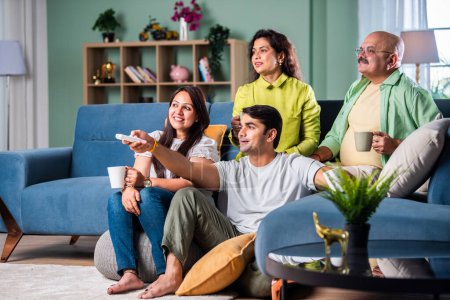 Téléchargez les photos : Famille indienne de quatre sur canapé, absorbé par regarder la télévision, changer de chaînes avec télécommande - en image libre de droit