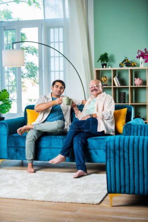 Photo for Happy Indian father and son having tea or coffee while sitting on sofa in living room - Royalty Free Image