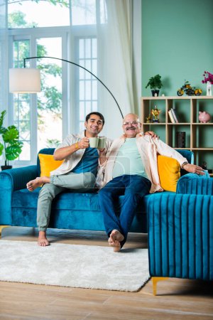 Foto de Padre e hijo indios felices tomando té o café mientras están sentados en el sofá en la sala de estar - Imagen libre de derechos