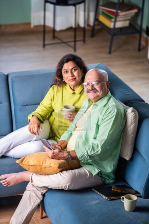 Téléchargez les photos : Heureux couple indien âgé utilisant Smartphone naviguant sur Internet ensemble assis sur le canapé à la maison - en image libre de droit