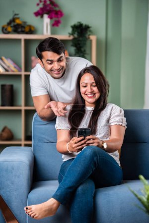 Photo for Happy and beautiful Indian Asian young couple using cellphone while sitting on sofa or couch - Royalty Free Image