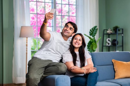 Téléchargez les photos : Heureux et beau couple asiatique indien jeune en utilisant un téléphone portable tout en étant assis sur le canapé ou le canapé - en image libre de droit