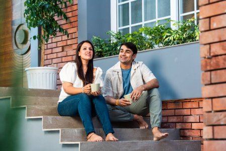 Téléchargez les photos : Heureux jeune couple indien prendre le thé ou le café ensemble tout en étant assis sur les escaliers à l'extérieur - en image libre de droit