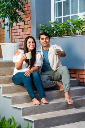 Téléchargez les photos : Heureux jeune couple indien prendre le thé ou le café ensemble tout en étant assis sur les escaliers à l'extérieur - en image libre de droit