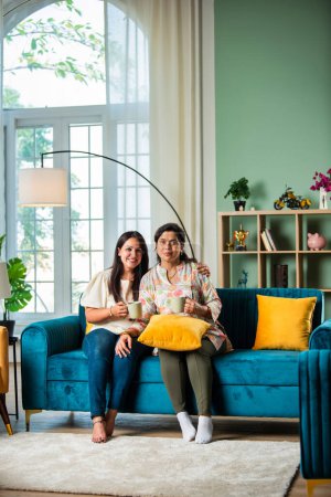 Téléchargez les photos : Mère indienne mature avec fille adulte se détendre sur le canapé prendre un café à la maison - en image libre de droit