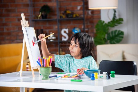 Photo for Indian little Boy Painting On Easel or on paper using brush and paint - Royalty Free Image