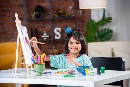 Photo for Indian little Boy Painting On Easel or on paper using brush and paint - Royalty Free Image