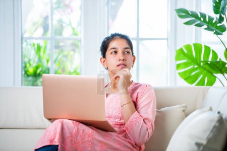 Téléchargez les photos : Indien jeune femme avec un ordinateur portable canapé assis dans un salon moderne - en image libre de droit