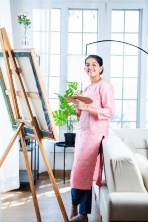 Photo for Cheerful Indian young artist girl painting picture at easel in living room - Royalty Free Image