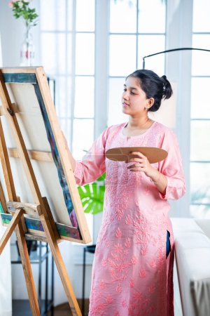 Photo for Cheerful Indian young artist girl painting picture at easel in living room - Royalty Free Image