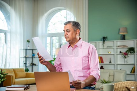 Foto de Hombre de mediana edad indio que trabaja en el ordenador portátil, teléfono inteligente con documentos que tienen café - Imagen libre de derechos