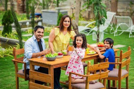 Photo for Indian young family of four eating meal on dining table in the garden - Royalty Free Image