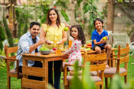 Téléchargez les photos : Jeune famille indienne de quatre personnes mangeant sur une table à manger dans le jardin - en image libre de droit