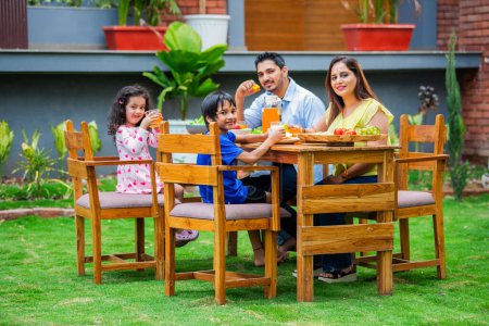 Téléchargez les photos : Jeune famille indienne de quatre personnes mangeant sur une table à manger dans le jardin - en image libre de droit