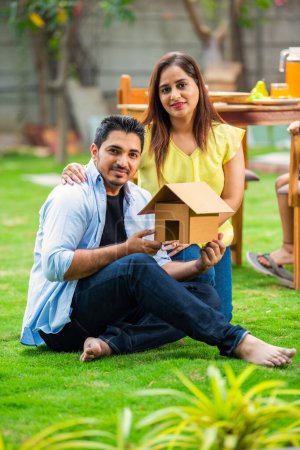 Téléchargez les photos : Portrait de jeune couple indien tenant modèle de maison tout en étant assis sur l'herbe verte à l'extérieur - en image libre de droit